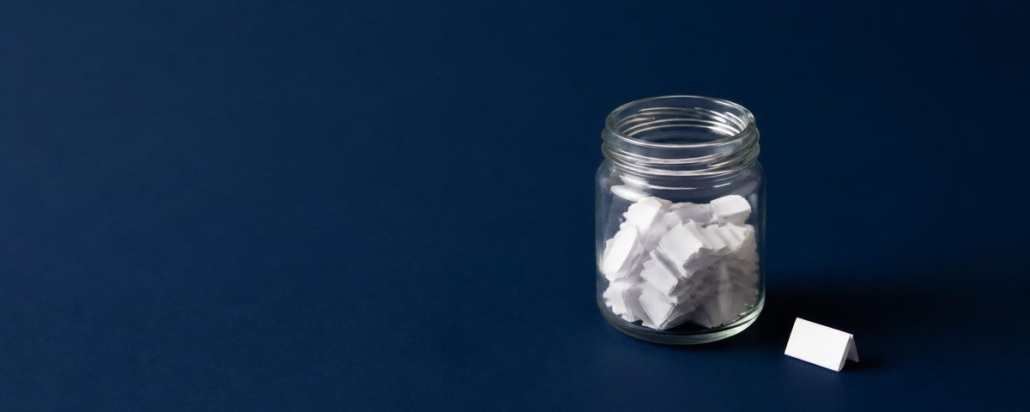 Small glass jar partly filled with folded white paper slips on a navy background, with one more folded slip beside it waiting to be added