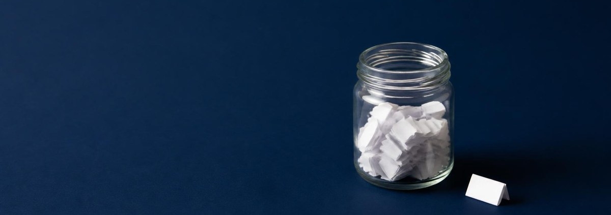 Small glass jar partly filled with folded white paper slips on a navy background, with one more folded slip beside it waiting to be added