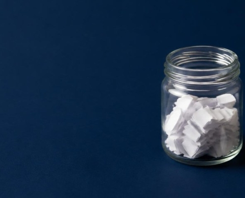 Small glass jar partly filled with folded white paper slips on a navy background, with one more folded slip beside it waiting to be added
